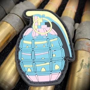 Puede incluir: Un parche colorido con forma de granada. El diseño presenta un cuerpo azul claro con detalles en rosa y amarillo, delineado en negro. El parche está sobre un fondo de casquillos de bala de latón.
