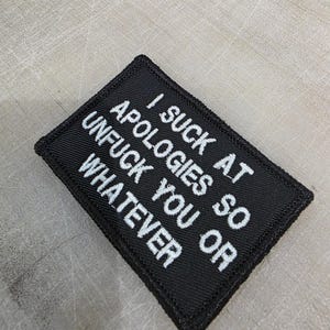 May include: Black rectangular patch with white embroidered text. The text reads "I SUCK AT APOLOGIES SO UNFU*K YOU OR WHATEVER". The patch has a black border and is likely made of fabric.