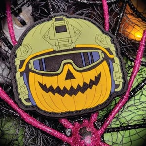 Puede incluir: Un parche temático de Halloween con una calabaza con un casco y gafas militares. La calabaza tiene una cara tallada con una boca dentada. El casco es verde oliva, las gafas son negras y azules, y la calabaza es amarilla.