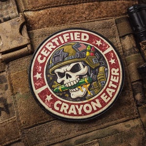 Puede incluir: Un parche redondo con el texto "CERTIFIED CRAYON EATER" en un diseño rojo y blanco. Presenta una calavera con un casco militar y sosteniendo un crayón verde. El parche está adherido a un chaleco de camuflaje.