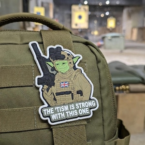 Peut inclure: Un écusson représentant un extraterrestre vert en tenue militaire, tenant un fusil. L'écusson comprend le texte "THE 'TISM IS STRONG WITH THIS ONE" et un drapeau britannique. L'écusson est fixé à un sac à dos vert.