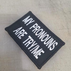 以下が含まれることがあります： 「MY PRONOUNS ARE TRYME」と白い刺繍が施された黒い長方形のパッチ。 パッチには黒い縁取りがあり、衣服やアクセサリーに縫い付けたり、アイロンをかけたりするように設計されています。