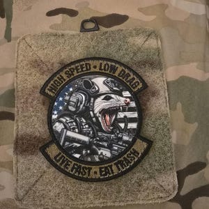 Pode incluir: Remendo circular com o texto "HIGH SPEED - LOW DRAG" e "LIVE FAST - EAT TRASH". O remendo apresenta uma ilustração detalhada de um gambá usando equipamento tático e segurando uma arma, com um design da bandeira americana no fundo.