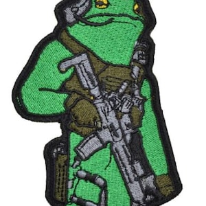 Puede incluir: Parche bordado que representa una rana verde con uniforme militar, sosteniendo un rifle. El parche tiene un borde negro y detalles en verde, gris y marrón. La rana lleva una capucha.
