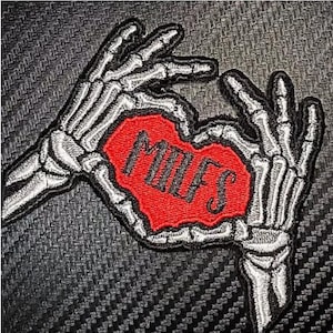 May include: Embroidered patch featuring two skeletal hands forming a heart shape around a red heart. The heart contains the word "MILFS" in black gothic lettering. The patch has a black and gray textured background.
