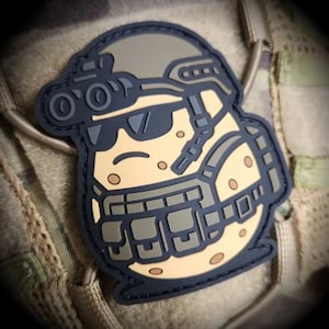 May include: A tactical patch featuring a cartoon potato character wearing a helmet, sunglasses, and tactical gear. The patch is primarily tan, gray, and black, with a dark blue border. The potato has a smiling expression.