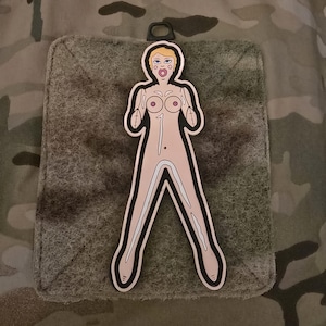 May include: A pink and white inflatable doll patch with a black outline. The doll has blonde hair, a pink face, and a surprised expression. The doll is wearing a pink dress and has a black strap on its back.
