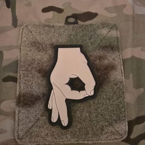 Könnte beinhalten: Ein schwarz-weißes Patch mit einer Hand, die die "OK"-Geste macht. Die Hand ist schwarz umrandet und mit hellrosa gefüllt.