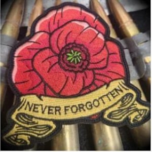 Può includere: Toppa ricamata raffigurante un fiore di papavero rosso con centro verde e contorno nero. Uno striscione giallo sottostante recita "NEVER FORGOTTEN" in testo nero. La toppa è posta su uno sfondo di bossoli di ottone.