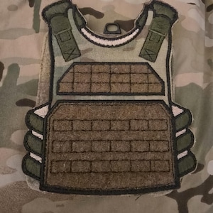 May include: A green and brown embroidered patch depicting a tactical vest with a front plate and shoulder straps.