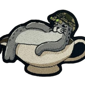 Puede incluir: Parche bordado con un leopardo de las nieves de dibujos animados que lleva un sombrero de camuflaje con gafas de visión nocturna, descansando en una taza de té. El leopardo es gris y blanco, la taza es beige con un asa y borde negros. El parche tiene un borde negro.