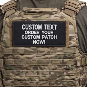 May include: A camouflage tactical vest with a black rectangular patch. The patch features white text that reads "CUSTOM TEXT ORDER YOUR CUSTOM PATCH NOW!". The vest has multiple straps and a modular design.