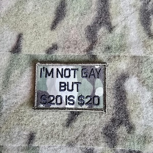 Op de afbeelding: Rechthoekige patch met de tekst "I'M NOT GAY BUT $20 IS $20" in zwarte letters. De patch heeft een camouflagepatroon achtergrond en een zilveren rand. De patch bevindt zich op een camouflage stof.