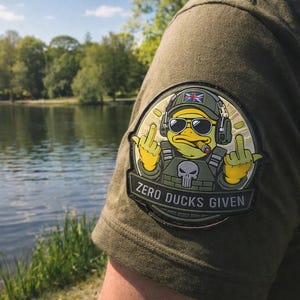 May include: Olive green t-shirt with a patch featuring a cartoon duck wearing sunglasses, a military helmet with a British flag, and headphones. The duck is giving the middle finger and smoking a cigar. The patch reads "ZERO DUCKS GIVEN."