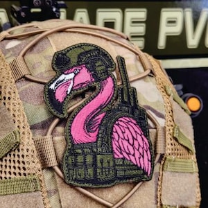 May include: An embroidered patch featuring a pink flamingo in military gear, including a helmet and vest, with a radio. The patch is in black, green, and pink, attached to a camouflage helmet.