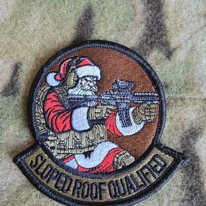 May include: A brown and tan embroidered patch with a cartoon image of Santa Claus holding a rifle. The text "Sloped Roof Qualified" is written in a black circle around the image.