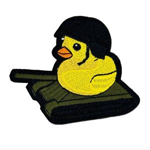 Puede incluir: Parche bordado con un pato amarillo con un casco negro, sentado en un tanque verde y negro. El pato tiene un pico naranja y ojos negros. El parche tiene un borde negro y está sobre un fondo blanco.