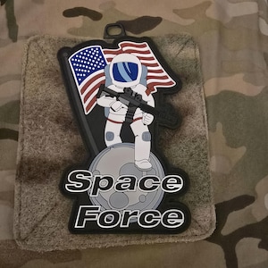 Könnte beinhalten: Ein schwarz-weißes Patch mit einem Astronauten, der ein Gewehr hält, auf dem Mond steht und eine amerikanische Flagge hinter sich hat. Der Text "Space Force" steht unter dem Astronauten.