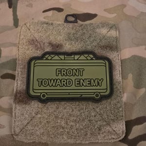 May include: An olive green patch with the text "FRONT TOWARD ENEMY" in white capital letters. The patch has a black border and is attached to a camouflage fabric. The patch is rectangular in shape.