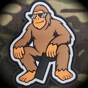 May include: A brown cartoon ape patch with orange hands and feet, wearing pixelated sunglasses. The ape is sitting down with a black rectangle in front of it. The patch has a white border and a black outline.