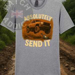 May include: A grey t-shirt with a graphic of a lifted truck and the text "ABSOLUTELY SEND IT" in yellow. The truck is illustrated in brown and orange, with a blue exhaust pipe. The shirt has a crew neck and short sleeves.