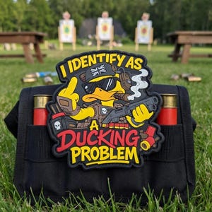Può includere: Pochette tattica nera con due bossoli rossi per fucile. Una toppa sulla pochette raffigura un'anatra dei cartoni animati con occhiali da sole, cuffie e che tiene in mano una pistola. La toppa recita "I IDENTIFY AS A DUCKING PROBLEM."