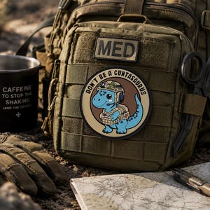 Può includere: Uno zaino tattico verde oliva con una toppa "MED" e una toppa circolare con un dinosauro blu in tenuta militare. Una tazza nera con la scritta "CAFFEINE TO STOP THE SHAKING (AND THE CRYING)" è accanto allo zaino.