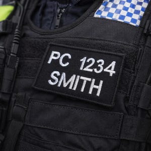 May include: Close-up of a black police vest with the text "PC 1234 SMITH" embroidered in white. A handheld radio and a yellow and blue checkered patch are also visible. The vest is part of a law enforcement uniform.