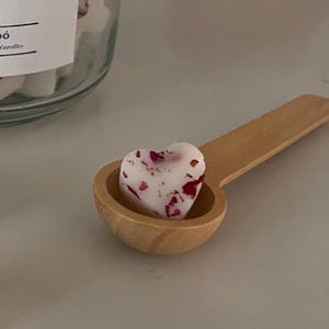 May include: A wooden spoon holds a heart-shaped wax melt with red and white flecks. The wax melt is in a jar labelled "Gel Wax Melts" and "Candles".