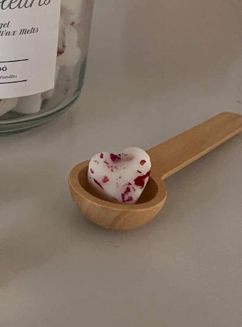 May include: A wooden spoon holds a heart-shaped wax melt with red and white flecks. The wax melt is in a jar labelled "Gel Wax Melts" and "Candles".