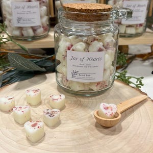 May include: A glass jar filled with white heart-shaped soy wax melts with red flecks. The jar has a cork lid and a label that reads "Jar of Hearts" with the text "Fresh Lemon Handmade Soy Wax Melts" and "Bea & Boo". There are several white heart-shaped wax melts on a wooden surface.