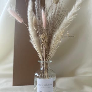 May include: A clear glass bottle with a white label that reads "Pear & Elderflower" and a bouquet of dried pampas grass in shades of white and pink.
