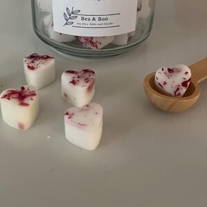 May include: A close-up of a wooden spoon holding a heart-shaped wax melt. The wax melt is white with pink and red flecks. There are several other heart-shaped wax melts on a white surface. The wax melts are labelled "Bea & Boo" soy wax melts and candles.