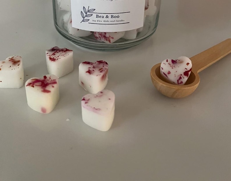 May include: A close-up of a wooden spoon holding a heart-shaped wax melt. The wax melt is white with pink and red flecks. There are several other heart-shaped wax melts on a white surface. The wax melts are labelled "Bea & Boo" soy wax melts and candles.