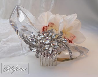 Puede incluir: Una zapatilla de cristal transparente con un peine plateado adornado con cristales transparentes y pedrería. Una flor blanca y moteada de rojo descansa detrás del peine. La palabra "Estefania" está en la esquina inferior izquierda.