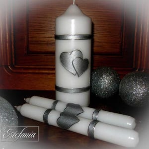 May include: White pillar candle and taper candles with silver heart and band accents. Two glittery silver decorative spheres are in the background. The candles are arranged on a wooden surface. The image includes the text "Estefania".