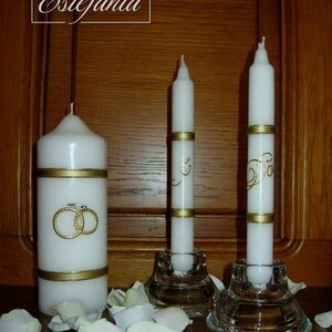 Puede incluir: Velas blancas con detalles dorados y diseños de anillos de boda. Las velas están en candelabros de vidrio, rodeadas de pétalos de flores blancas. Se ve la palabra "Estefania".