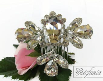 Puede incluir: Un peine para el cabello plateado adornado con grandes y brillantes cristales transparentes. El peine presenta un grupo central de cristales con detalles en forma de ala y un cristal colgante en forma de lágrima. Una rosa rosa y hojas verdes en el fondo.