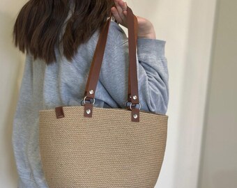 May include: A tan woven tote bag with brown leather straps and silver hardware. The bag has a wide, rectangular shape and is made of a thick, textured material.