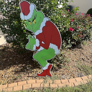 May include: A green Grinch character wearing a red Santa suit and a white hat. The Grinch is standing on a bed of brown gravel with a brick border.