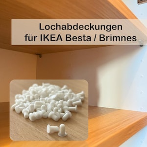 Könnte beinhalten: Weiße Kunststoff-Lochabdeckungen für IKEA Besta / Brimnes Möbel. Das Bild zeigt einen Haufen der Abdeckungen auf einer Holzoberfläche, mit ein paar einzelnen Abdeckungen im Vordergrund. Der Text im Bild lautet "Lochabdeckungen für IKEA Besta / Brimnes."