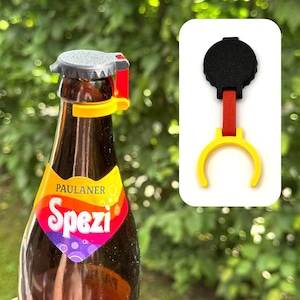 K&ouml;nnte beinhalten: Ein Flaschen&ouml;ffner-Aufsatz mit einem schwarzen runden Oberteil, einem roten vertikalen Verbindungsst&uuml;ck und einem gelben Ringfu&szlig;. Das Ger&auml;t ist am Hals einer braunen Glasflasche mit dem bunten Etikett Paulaner Spezi befestigt.
