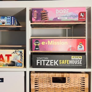 Könnte beinhalten: Ein weißes Regal zeigt Brettspiele. Zu den Kartons gehören "SKY TEAM", "HEAT", "DORF ROMANTIK SAKURA", "e+Mission" und "SEBASTIAN FITZEK SAFEHOUSE". Unten befindet sich ein Weidenkorb. Die Spiele stehen auf Holzregalen.