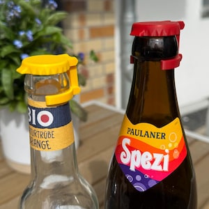 Könnte beinhalten: Zwei Glasflaschen mit bunten Etiketten, eine mit der Aufschrift "BIO Naturtrübe Orange" und die andere mit "PAULANER Spezi". Jede Flasche hat einen Kunststoffverschluss in Gelb bzw. Rot. Die Flaschen stehen auf einer Holzoberfläche.