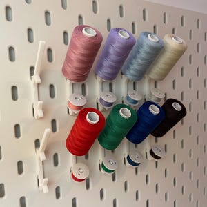 Könnte beinhalten: Eine Sammlung farbiger Garnrollen und Spulen, die auf einer weißen Lochwand ausgestellt sind. Die Garnfarben umfassen Rosa, Lila, Blau, Beige, Rot, Grün, Dunkelblau und Braun. Die Spulen passen zu den Garnfarben.