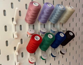 Könnte beinhalten: Eine Sammlung farbiger Garnrollen und Spulen, die auf einer weißen Lochwand ausgestellt sind. Die Garnfarben umfassen Rosa, Lila, Blau, Beige, Rot, Grün, Dunkelblau und Braun. Die Spulen passen zu den Garnfarben.