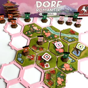 Könnte beinhalten: Ein Brettspiel mit einem rosa sechseckigen Spielbrett und farbenfrohen Geländefeldern. Holzmarker in Braun, Grün und Rosa sind auf dem Spielbrett platziert. Die Spielbox mit dem Titel "DORF ROMANTIK" und einer Pagodenillustration ist sichtbar.