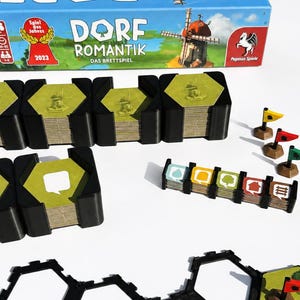 Könnte beinhalten: Eine Dorfromantik Brettspielschachtel mit einer Illustration von blauem Himmel und Windmühle, dazu mehrere Stapel sechseckiger Spielplättchen in schwarzen Halterungen, eine kleine Reihe markierter Plättchen und drei kleine Flaggen.