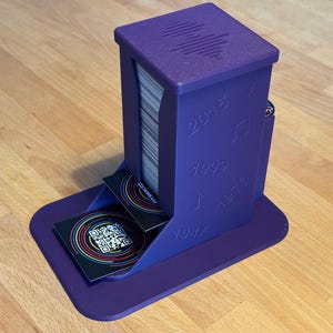 Könnte beinhalten: Ein lila Kartenhalter mit einem Stapel Karten. Der Halter hat die Jahre 1973, 1987, 1999 und 2015 sowie Notensymbole auf der Seite geprägt. Zwei Karten mit farbenfrohen kreisförmigen Designs und QR-Codes sind sichtbar.