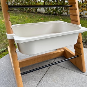 Könnte beinhalten: Ein hölzerner Hochstuhl mit einem weißen Tablett. Der Stuhl hat eine natürliche Holzoberfläche und schwarze Metallstangen. Das Tablett ist rechteckig und scheint aus Kunststoff zu sein. Der Hochstuhl ist für ein Baby oder Kleinkind konzipiert.